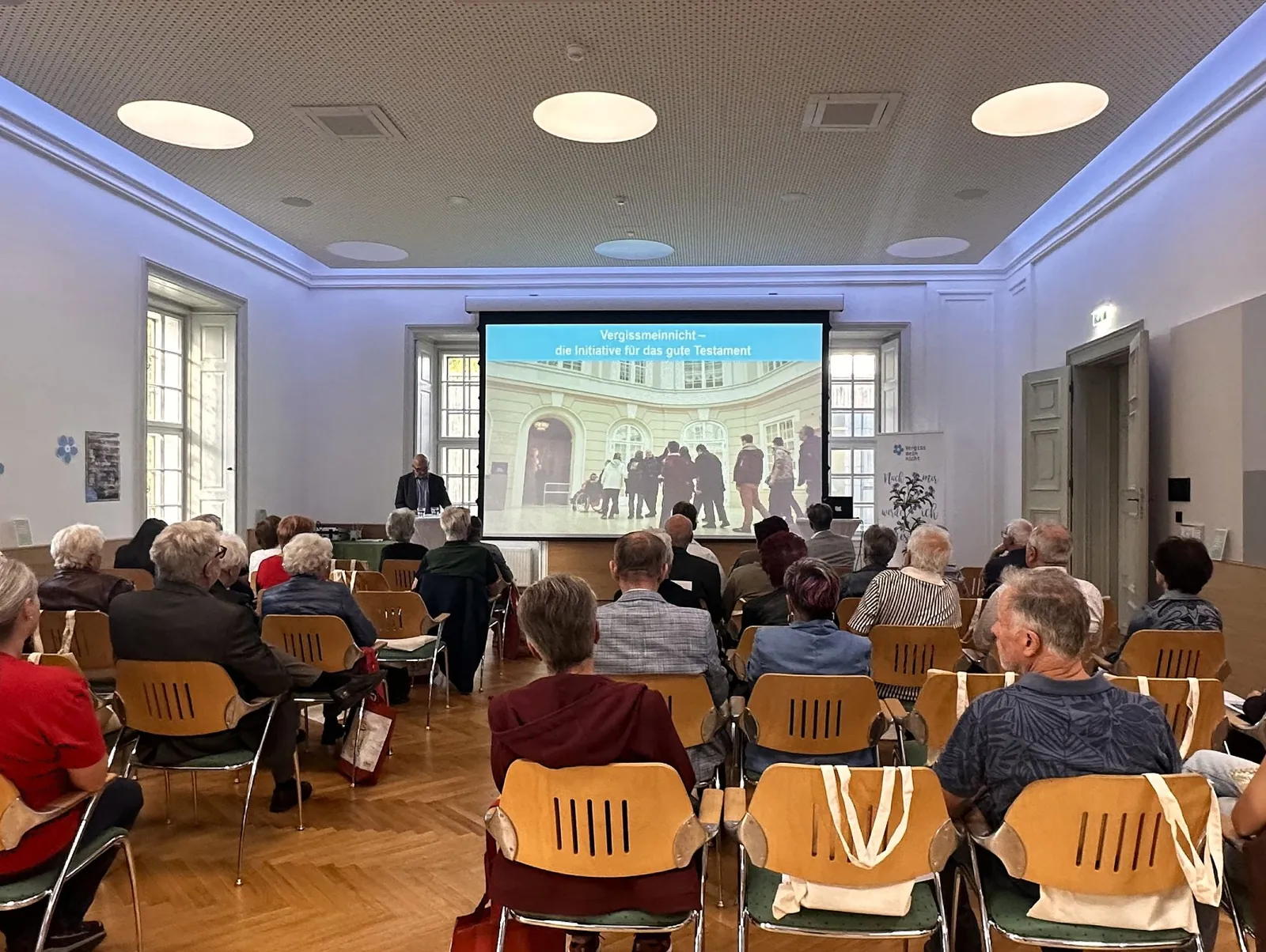 Publikum in Sesselreihen vor einer Leinwand mit der Aufschrift Vergissmeinnicht - die Initiative für das gute Testament